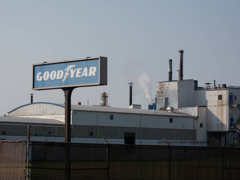 The Goodyear chemical plant is pictured. In front of it is a Goodyear logo sign, which is blue with white text that reads "Good," then an image of a stepping shoe with wings, and then the word "year." The factory behind it has a dark wire fence surrounding it, and is white and grey. The factory has a white puff of smoke coming out of the top of it. Rust runs down the side of some of the outer walls.