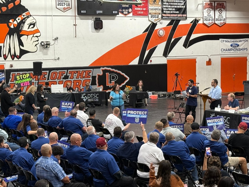 A view inside a high school gymnasium, with orange and black paint on the walls; many attendees of a public meeting are wearing blue shirts, and some hold signs saying “Protect Teamster Jobs” while a woman in a light blue shirt speaks to the group.