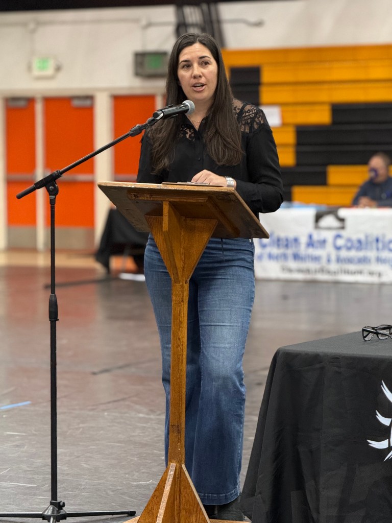 A photo of a woman with brown hair, wearing a black shirt and jeans, standing behind a small wooden podium, with a standing microphone pointed in her direction.