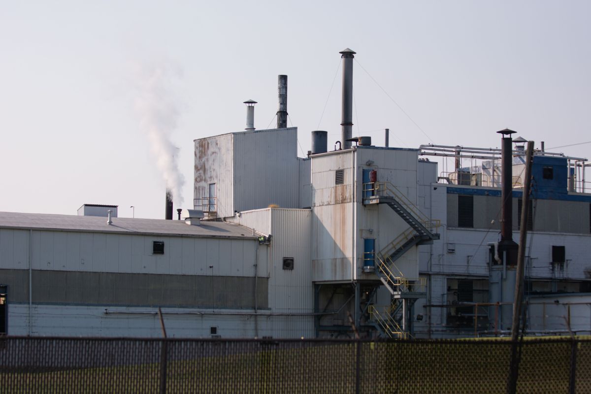 The Goodyear chemical plant is pictured. The factory has a dark wire fence surrounding it, and is white and grey. The factory has a white puff of smoke coming out of the top of it. Rust runs down the side of some of the outer walls.