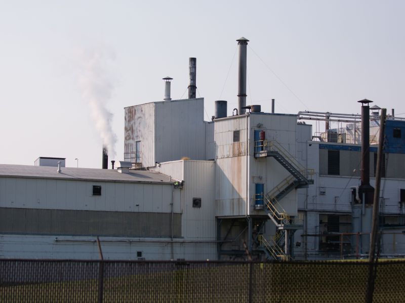 The Goodyear chemical plant is pictured. The factory has a dark wire fence surrounding it, and is white and grey. The factory has a white puff of smoke coming out of the top of it. Rust runs down the side of some of the outer walls.
